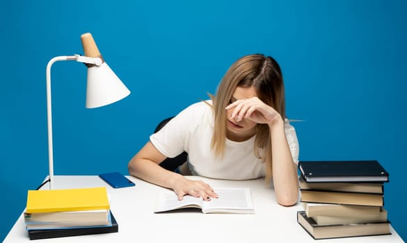 Tired young student woman in white casual clothes sitting at the table reading the book in library of university or college. Sitting and reading on blue background. Studying
