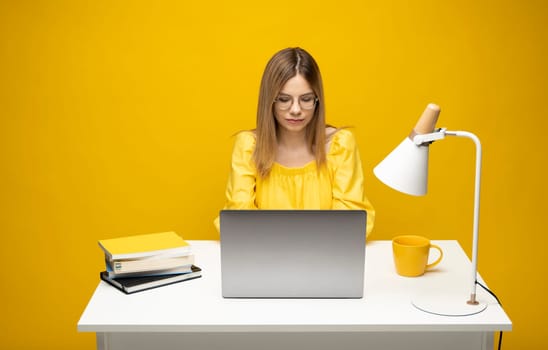 Portrait of a pretty young woman studying while sitting at the table with grey laptop computer, notebook. Smiling business woman working with a laptop isolated on a yellow background
