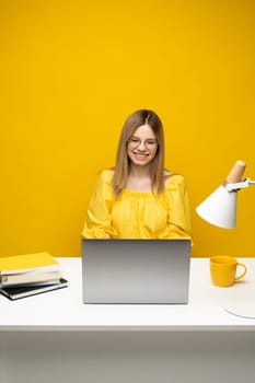 Excited young woman working with a grey laptop computer, notebook while sitting at the table with grey laptop. Smiling business woman or student received a good news isolated on a yellow background