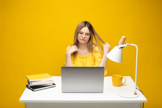 Portrait of Smiling pretty young woman studying while sitting at the table with grey laptop computer, notebook. Business woman working with a laptop isolated on a yellow background