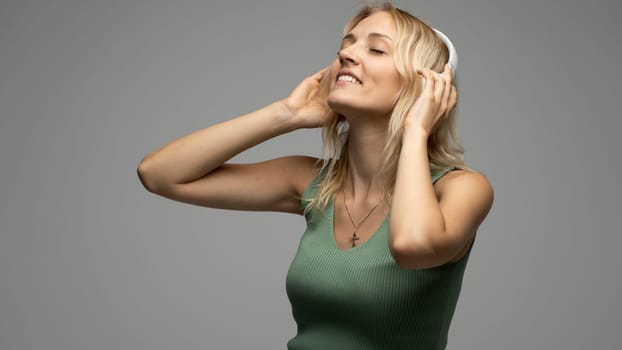 Beautiful attractive young blond woman wearing green t-shirt and glasses in white headphones listening music and smiling on grey background in studio. Relaxing and enjoying. Lifestyle