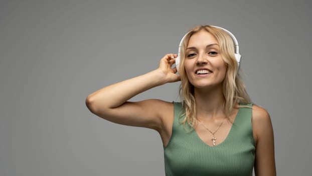 Beautiful attractive young blond woman wearing green t-shirt and glasses in white headphones listening music and smiling on grey background in studio. Relaxing and enjoying. Lifestyle