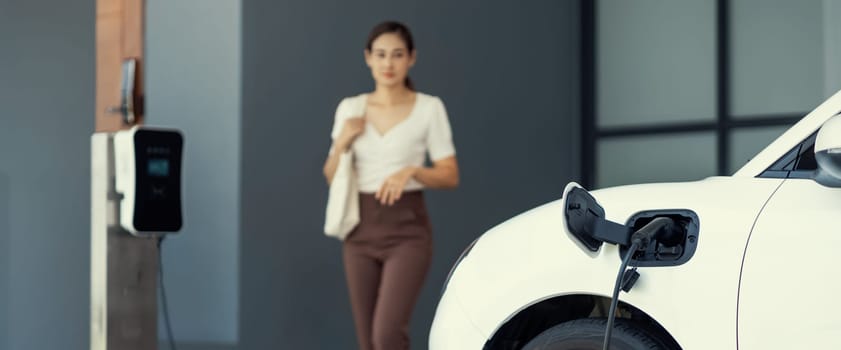 Focus image of electric vehicle recharging battery at home charging station with blurred woman walking in the background. Progressive concept of green energy technology applied in daily lifestyle.