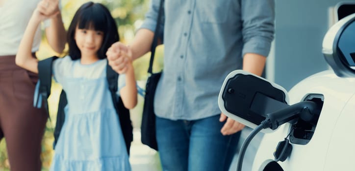 Focus closeup electric vehicle recharging battery from home electric charging station with blurred family in background. Renewable clean energy car for progressive eco awareness lifestyle concept.