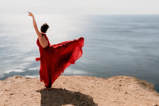 Side view a Young beautiful sensual woman in a red long dress posing on a rock high above the sea during sunrise. Girl on the nature on blue sky background. Fashion photo.