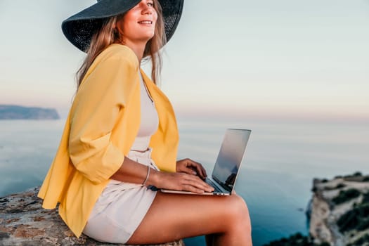 Successful business woman in yellow hat working on laptop by the sea. Pretty lady typing on computer at summer day outdoors. Freelance, travel and holidays concept.