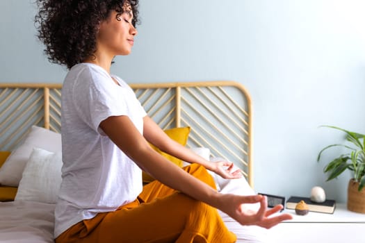 Side view of young multiracial woman relaxing on the bed. Meditating and breathing yoga exercises to calm mind. African American woman does meditation in bedroom. Wellness and meditation concept.