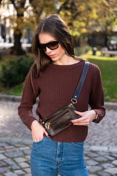 young brunette posing outside in a jeans and knitted sweater with a small brown belt bag