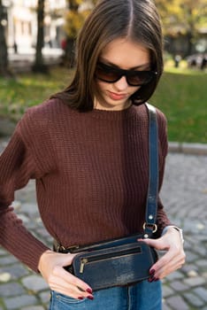 young brunette posing outside in a jeans and knitted sweater with a small brown belt bag