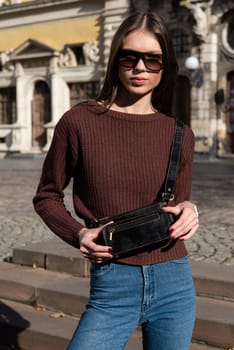 young brunette posing outside in a jeans and knitted sweater with a small brown belt bag