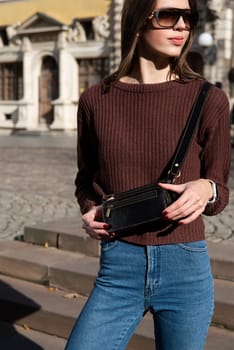 young brunette posing outside in a jeans and knitted sweater with a small brown belt bag