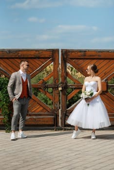 young couple bride in a white short dress and groom in a gray suit near the wooden rural gate of the estate