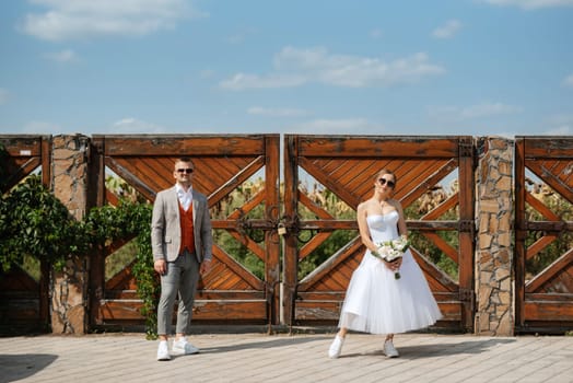 young couple bride in a white short dress and groom in a gray suit near the wooden rural gate of the estate