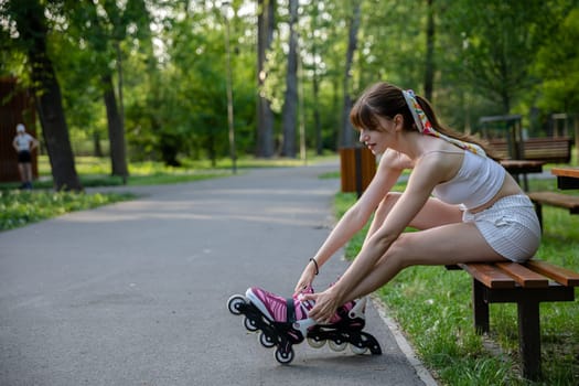 A woman improves the fastening of rollerblades while sitting on a bench. A girl puts on rollerblading sports equipment. In the background are fuzzy trees, lawns and an asphalt alley.