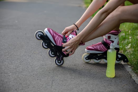 Skater girl straps rollerblades on her legs. Close-up of the legs of a young person in shorts. In the foreground a bottle of yellow isotonic drink standing on an asphalt alley.