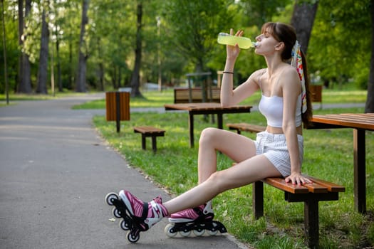 Spending time in a green city park. A young woman sits on a bench and drinks from a bottle. Replenishing electrolytes while playing sports. A hot day in nature.
