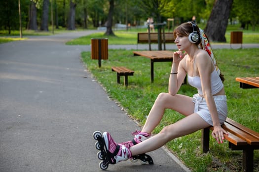 The girl is listening to music on wireless headphones. An athlete relaxes during training while sitting on a bench. Spending time in a green city park. A young woman dressed in sportswear.