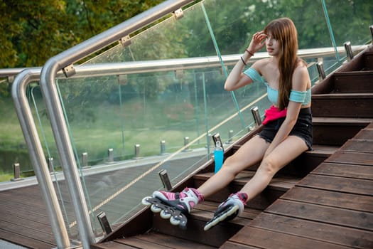 A girl on roller skates sits on a wooden staircase. The long-haired brunette is wearing a short summer sports outfit. She carries a hip bag on her belt. Next to the girl stands a bottle of blue drink.