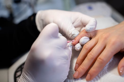 A manicurist paints a client's little finger with pink nail polish. The beautician's hands, wearing disposable latex gloves, are visible in the foreground.