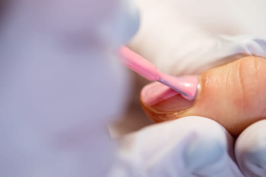 Application of hybrid varnish to the natural nail plate. Close-up on the hands of a beautician holding the finger of a client. A woman using the services of a beauty salon. Painting nails with hybrid method.