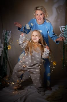 Cute mother and daughter in pajamas having fun in the room with Christmas garlands and white background. The tradition of decorating the house for holidays. Happy childhood and motherhood