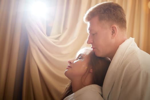 An adult couple of newlyweds in in white robes in a hotel room after the wedding. A guy and a girl, a man and a woman in beautiful room. The concept of hugs, love and care