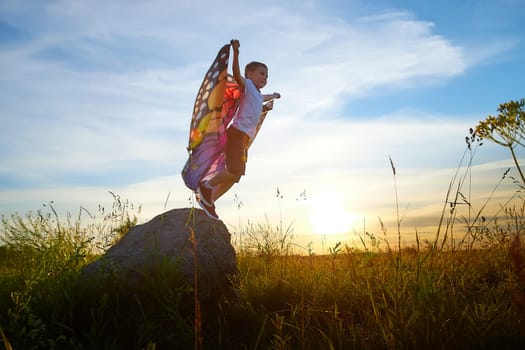 Handsome boy with bright butterfly wings having fun in meadow on natural landscape with grass and flowers on sunny summer day. Portrait of teenage guy in spring season outdoors on field