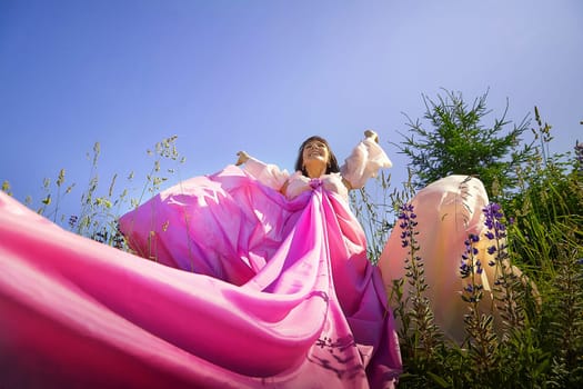 Beautiful girl in lush pink ball gown in green field during blooming of flowers and blue sky on background. Model posing on nature landscape as princess from fary tale