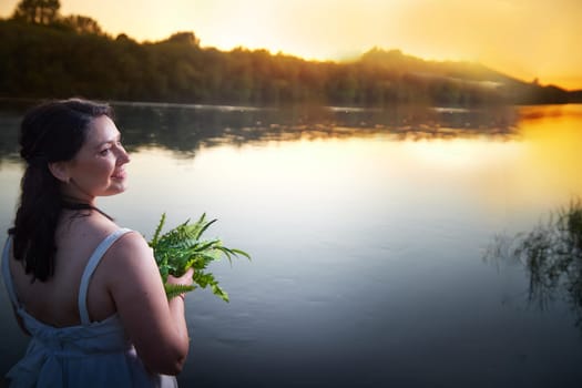 Slavic plump plump chubby girl in long white dress on the feast of Ivan Kupala with flowers and water in a river or lake on summer evening