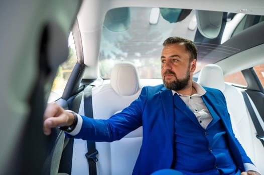 A caucasian man in a blue suit looks out the open window while sitting in the back seat of a car. Business class passenger