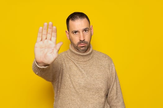 Hispanic man with a beard wearing a beige turtleneck in a serious attitude making the stop gesture with the palm of his hand isolated over yellow background