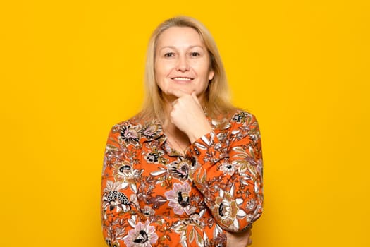 Closeup photo of amazing blonde lady looking into camera smiling, thinking deeply, creative person, arm on chin, patterned dress, isolated yellow color background