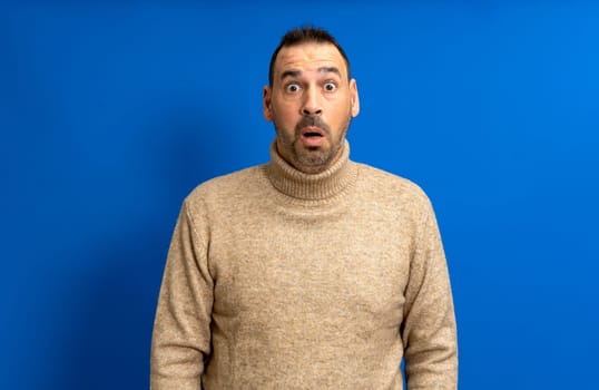 Bearded hispanic man wearing a turtleneck sweater with open mouth amazed and absorbed in something isolated over blue studio background