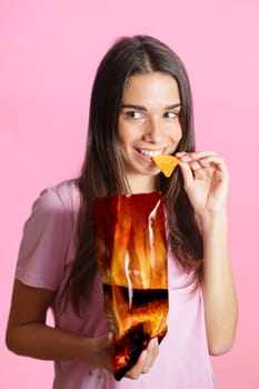 Smiling young female in casual clothes standing near bright pink background and biting crunchy potato chip while holding package