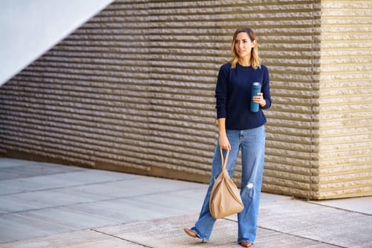 Full body woman in stylish jeans and sweater with bag and thermos looking away while standing on pavement outside modern building