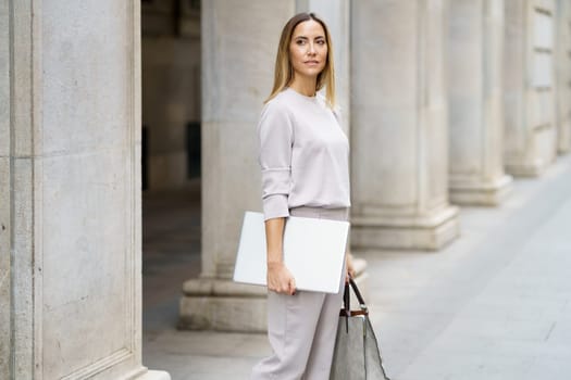 Serious self assured adult woman with blond hair in gray outfit standing on street near old building and holding laptop and handbag while looking away