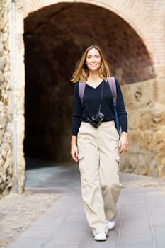 Full body of positive young female traveler with long hair in casual clothes and backpack, with photo camera hanging on neck smiling while walking in old city near arched passage