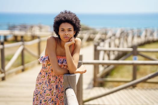 Young African American female tourist in ornamental dress with curly hair, leaning on hand and looking at camera while relaxing on boardwalk against sea and blue sky on summer day on resort