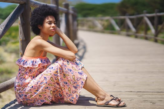 Side view of pensive ethnic female in summer dress and sandals with curly hair, leaning on hand and looking away while sitting on wooden path on sunny summer day