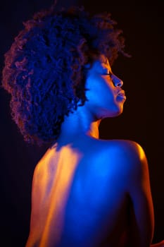Topless African American female with Afro hairstyle closing eyes under orange and blue neon light against black background