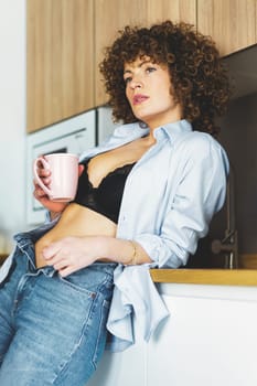 Curly haired female in black bra unbuttoned shirt and unzipped jeans leaning on counter while enjoying hot beverage