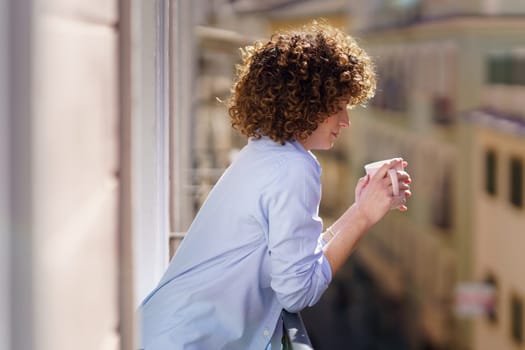 Side view of female with curly hair in blue shirt standing on small balcony and drinking hot beverage from cup against blurred background