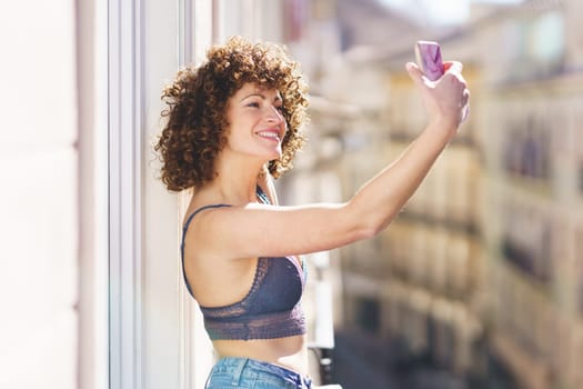 Cheerful young female in bra and jeans with curly hair taking selfie on smartphone while standing on balcony on sunny day