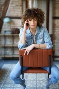 Positive young female with curly hair in jeans jacket sitting on leather chair and touching head in room with brick wall and tiled floor