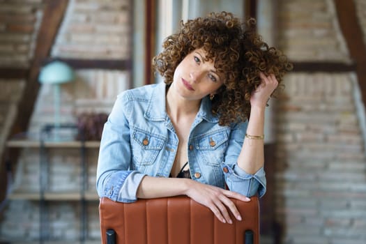 Serious young female in underwear and denim jacket sitting on leather chair and touching hair while looking at camera against blurred brick wall