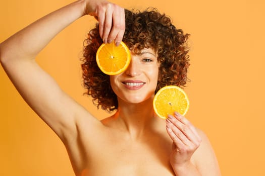 Happy woman with bare shoulders and curly hair, covering eye with slice of citrus and looking at camera with smile during skin care routine against orange background