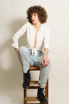 Full body of thoughtful female model with curly red hair in casual outfit chilling on wooden stool and looking away against white background
