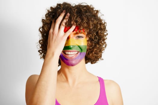 Happy curly haired female with painted in LGBT colors face covering eye looking at camera against white background equality and tolerance concept