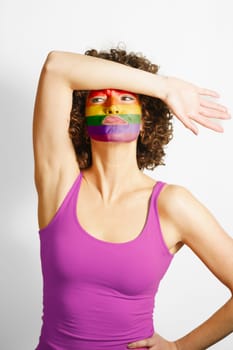 Young curly haired woman with hand on forehead and face painted in rainbow colors looking away while standing in studio solidarity and tolerance concept