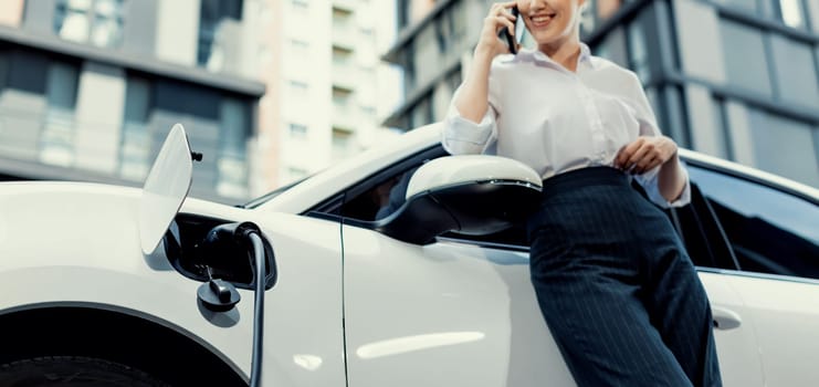 Closeup progressive businesswoman with smartphone, leaning electric car and charging station before driving around city center. Eco friendly rechargeable EV car powered by sustainable and clean energy